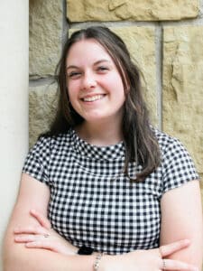 A smiling woman in a checkered shirt leaning against a stone wall at Howard Payne University. | HPU