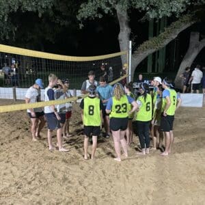 Two teams shaking hands over a volleyball net after a game at a night-time outdoor sand court at Howard Payne University. | HPU