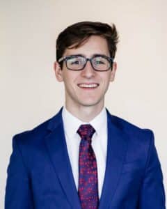 A smiling young man wearing glasses, a blue suit, and a red patterned tie from Howard Payne University. | HPU