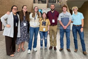A group of seven smiling people, four women and three men, standing indoors in a lobby, wearing name tags and casual to smart casual attire. | HPU