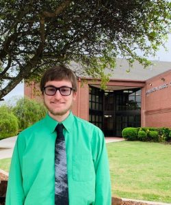 A smiling individual in a green shirt and black tie standing in front of a brick building at Howard Payne University with the sign "school of music". | HPU