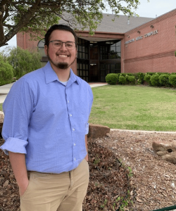 A smiling man in a blue shirt standing in front of a brick building at Howard Payne University, with a sign that reads "education service complex. | HPU