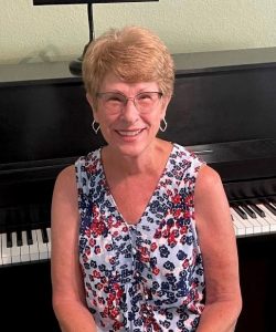 A smiling woman with short hair wearing glasses and a floral top sitting in front of a piano at Howard Payne University. | HPU