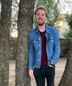 A man in a denim jacket and dark pants smiling between two trees on Howard Payne University campus. | HPU