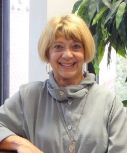 Smiling woman with short blonde hair, wearing a Howard Payne University gray outfit and necklace, seated indoors. | HPU