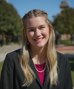 A smiling woman with long blonde hair wearing a black jacket over a purple top stands in front of Howard Payne University, with a park setting in the background. | HPU